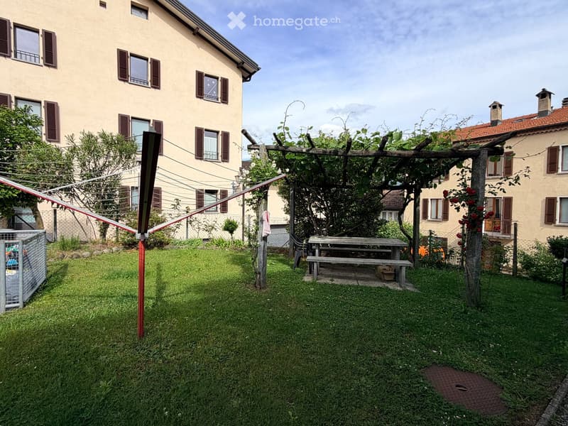 A well-maintained garden with a wooden pergola, picnic table, and surrounding residential buildings.