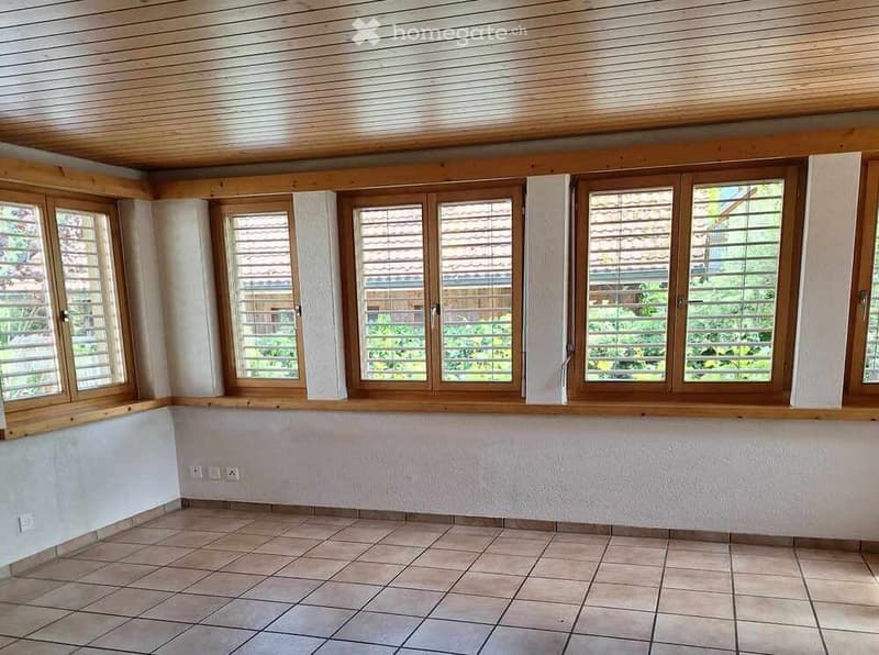 Empty room with tiled floor, wooden ceiling, and large windows with wooden frames and blinds.