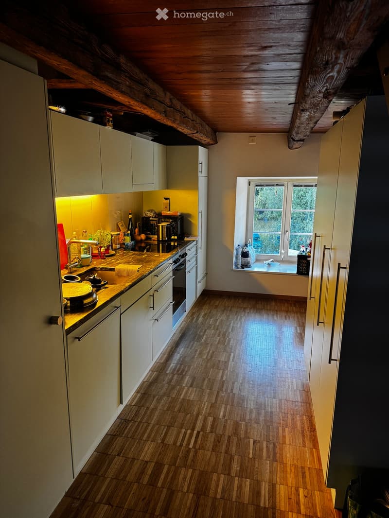 Modern kitchen with light-colored cabinets, wooden flooring, and a window overlooking greenery. The counter features a coffee maker, a bottle, a cup, and a bowl.