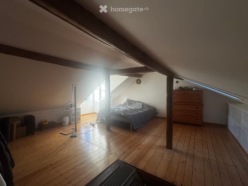 Cozy attic bedroom with wooden floors, slanted wooden ceiling beams, and large windows allowing natural light. Features a bed, nightstand, lamp, fan, and a small table.