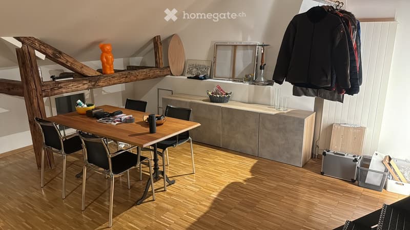 A modern, well-lit room with wooden flooring, a large wooden table surrounded by chairs, a built-in cabinet, and a shelf with decorative items and a suit hanging on the wall.