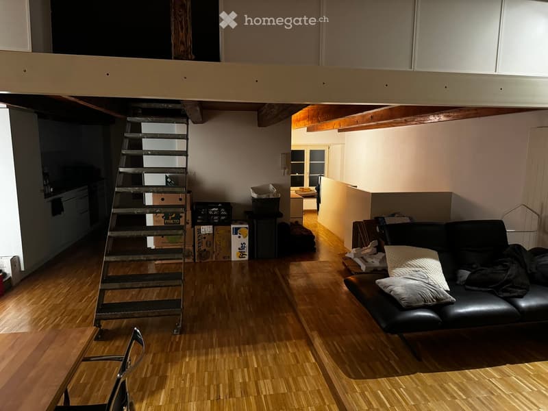 A furnished room with a black leather sofa, a wooden chair, a metal staircase leading to a loft area, and several cardboard boxes stacked against the wall.