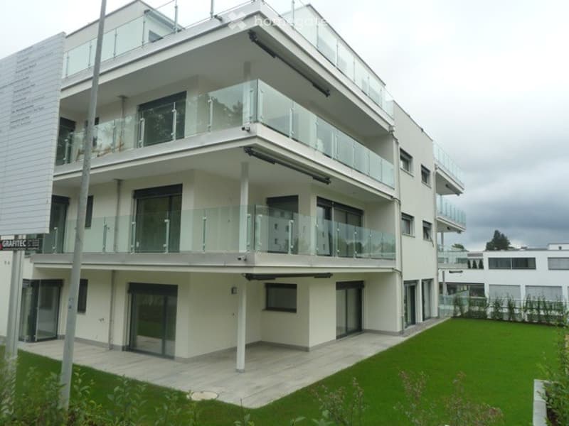 Modern three-story residential building with white facade, large glass balconies, and a well-maintained lawn in front.