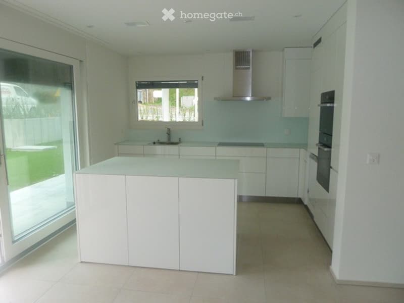 Modern, minimalist kitchen with white cabinets, a central island, and stainless steel appliances, including a range hood and oven. The space features large windows and a sleek, contemporary design.