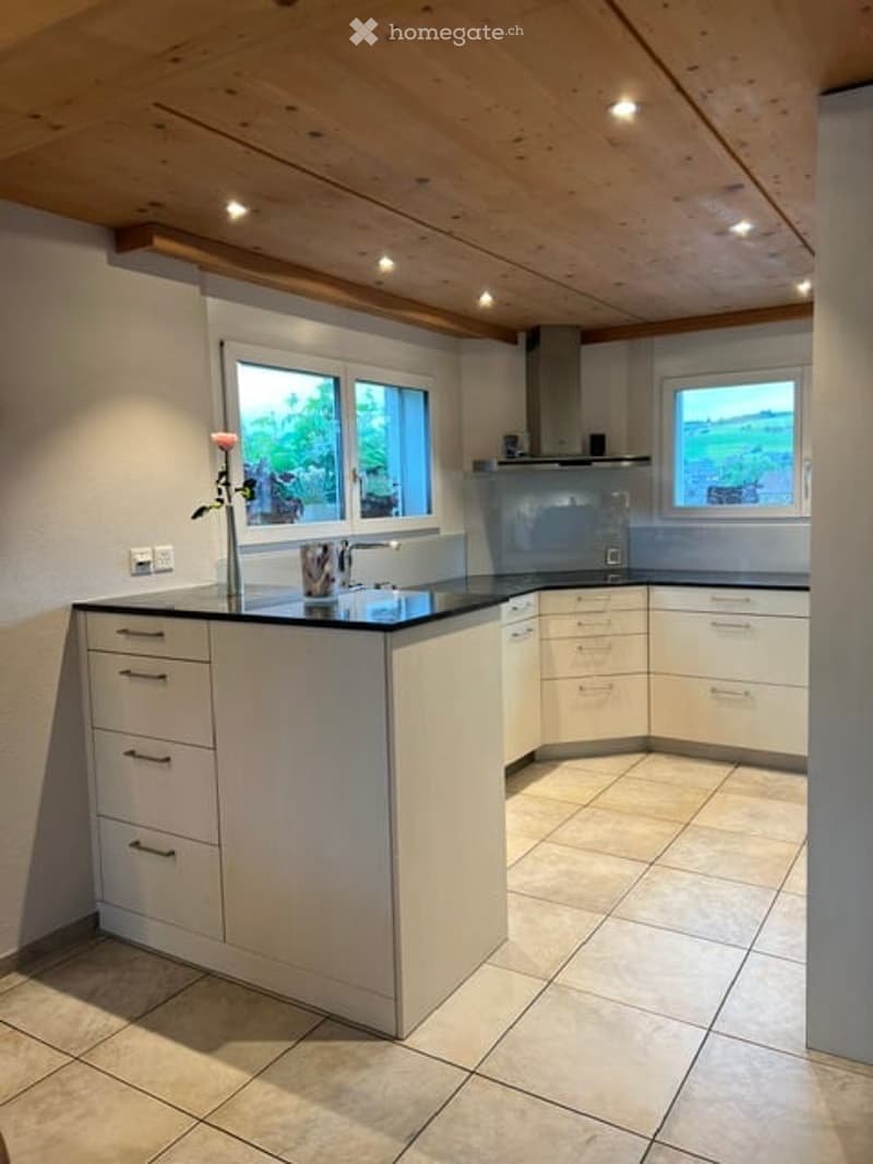 Modern kitchen with white cabinets, a central island, and a range hood. Features include a sink with a faucet, a flower vase on the island, and large windows offering a view.