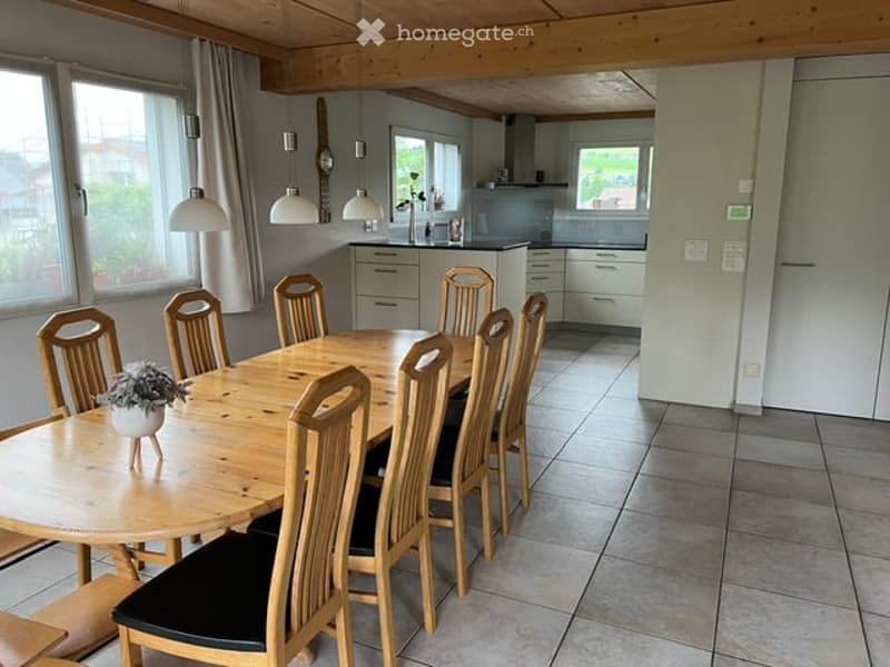 Bright, open-plan dining and kitchen area with wooden dining table, six chairs, pendant lights, and modern kitchen cabinets.
