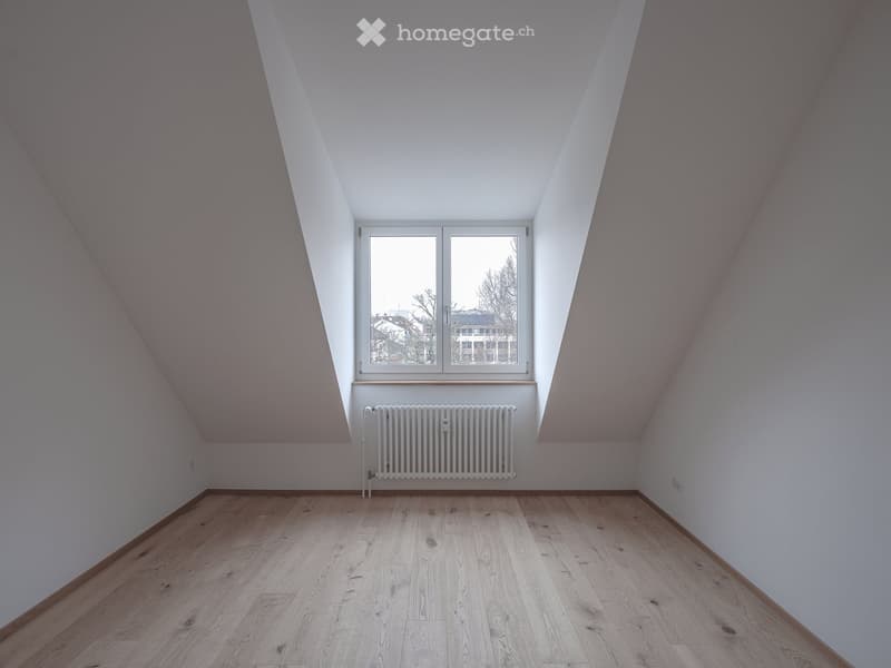 Empty attic room with wooden flooring, white walls, and a single window with a radiator below it.