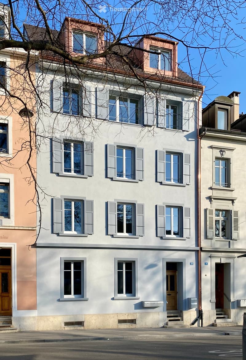 A three-story residential building with light gray walls, multiple windows with shutters, and a wooden front door.