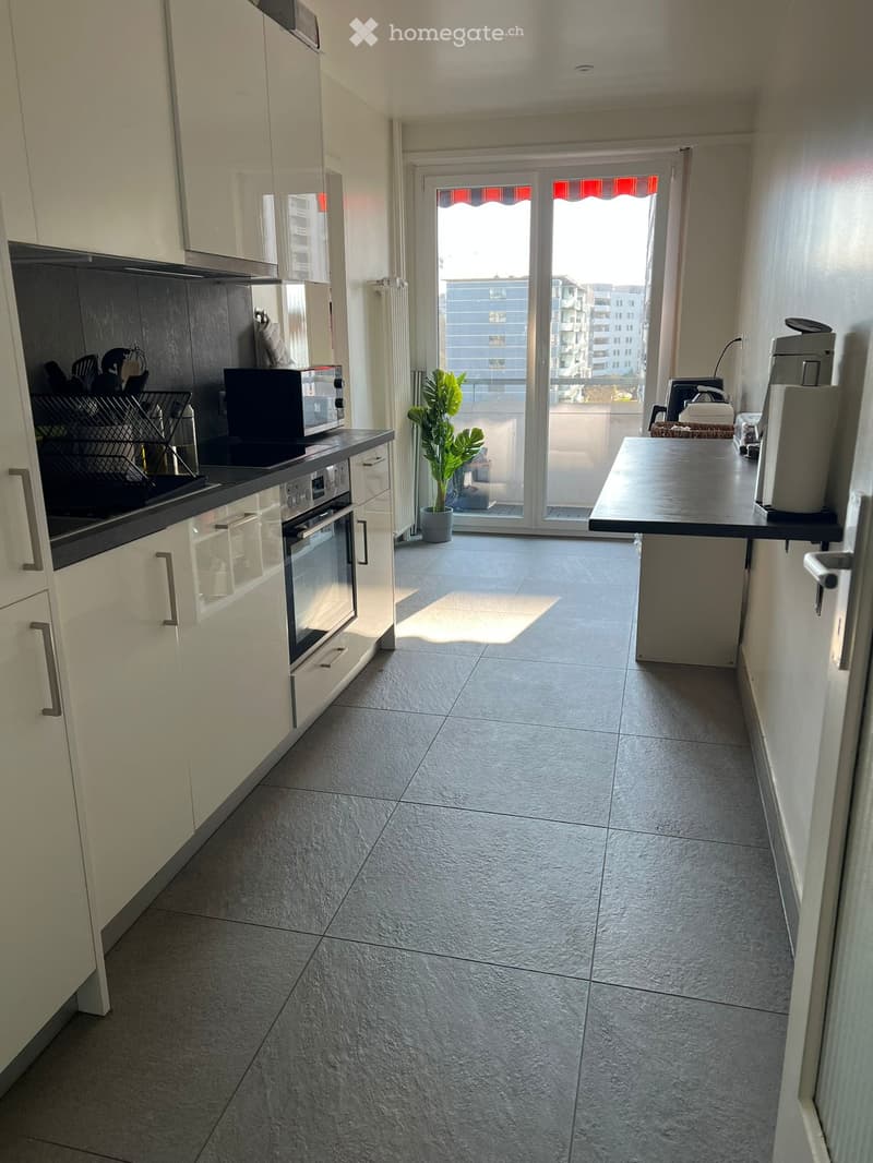 modern kitchen with white cabinets, gray tiled floor, built-in oven, microwave, and coffee machine, large windows with balcony access, potted plant, and city view