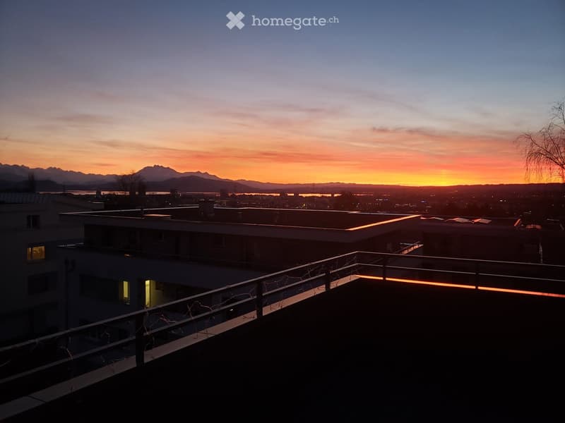 Balcony with a railing, overlooking a cityscape at sunset with mountains in the background.