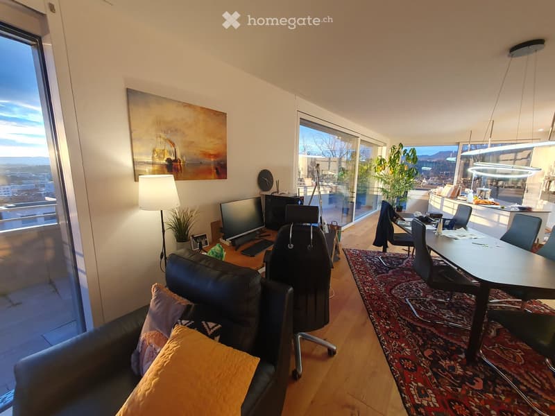 Modern living room with large windows offering a scenic view, featuring a black leather sofa with colorful cushions, a wooden coffee table with a screen and laptop, a large area rug, and a dining table with chairs.