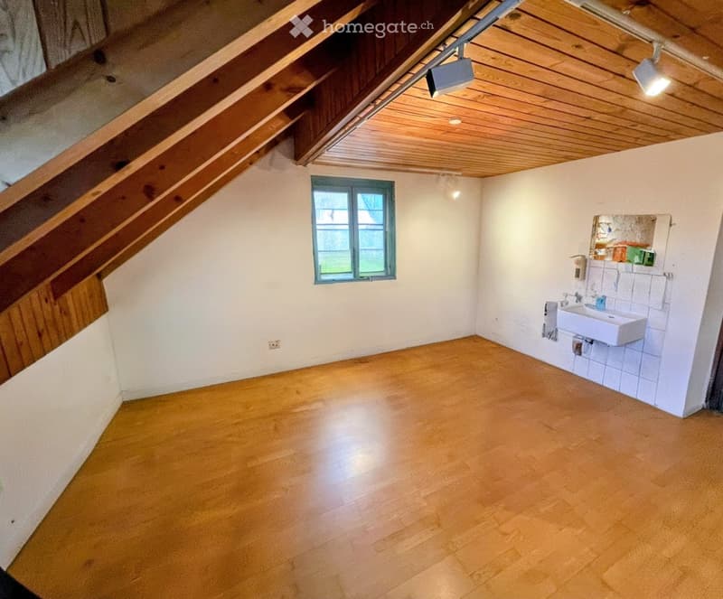 Empty attic space with wooden flooring, white walls, exposed wooden beams, and a small window. A small sink is installed in one corner.