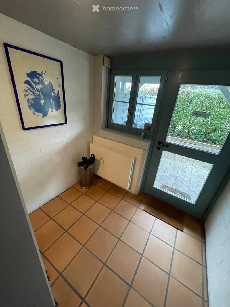 Hallway with tiled floor, white brick wall, radiator, umbrella stand, and a framed artwork on the wall. A door with glass panels leads outside.