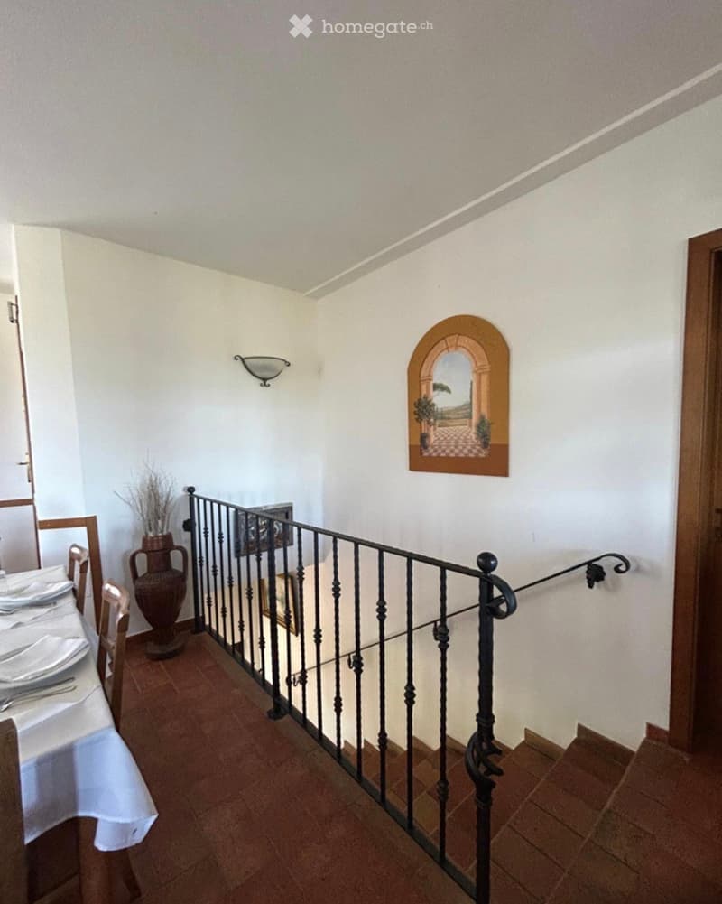 Staircase with black wrought iron railing, beige walls, terracotta tiled floor, decorative vase, and framed artwork on the wall.