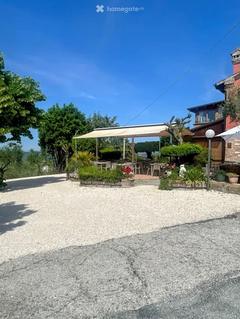 Outdoor seating area with tables, chairs, and an umbrella, surrounded by greenery and a gravel driveway.