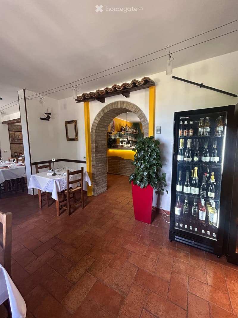 Restaurant interior with terracotta tiled floor, wooden tables and chairs, a large potted plant, and a refrigerator stocked with various bottles.