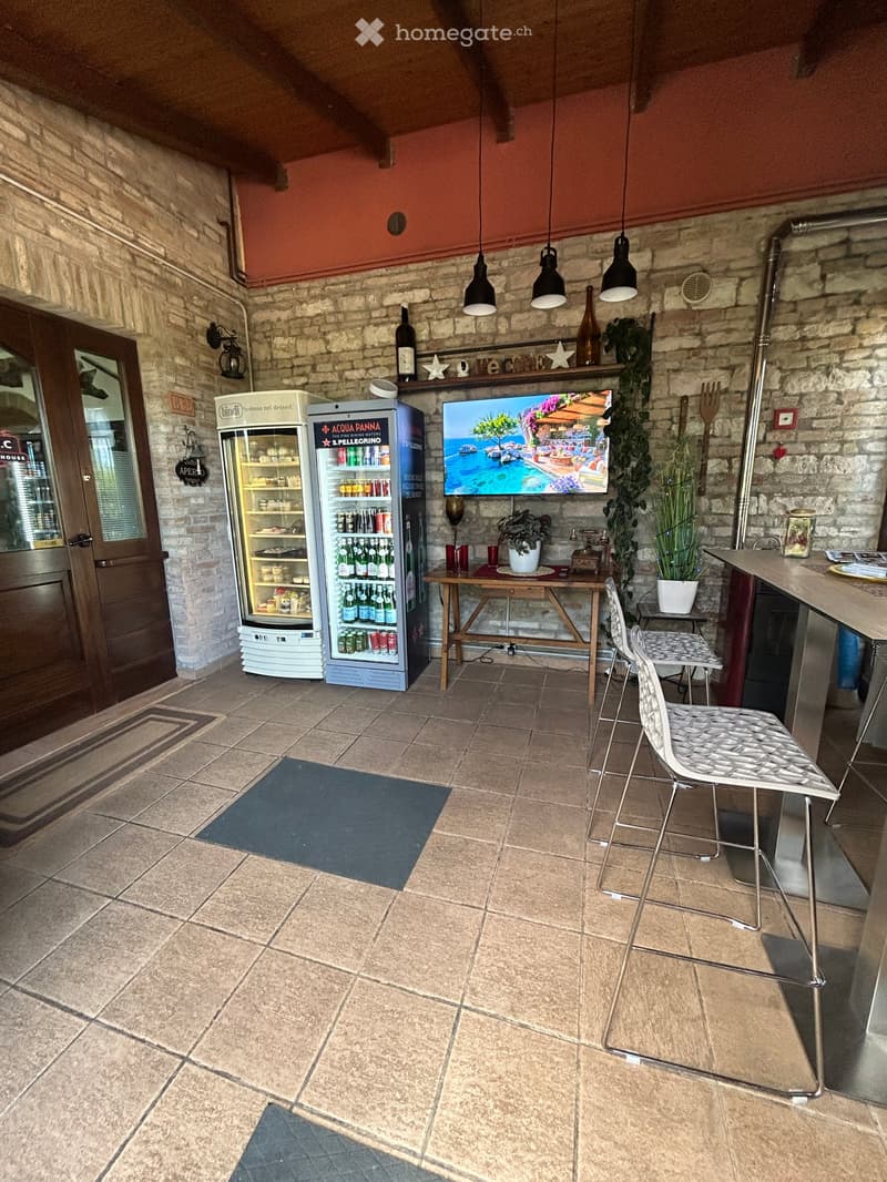 A cozy bar area with a refrigerator, a small table, chairs, a television, and potted plants. The space features exposed brick walls and a tiled floor.