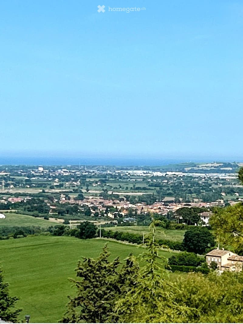 A panoramic view of a rural landscape with green fields, scattered houses, and a town in the distance under a clear sky.
