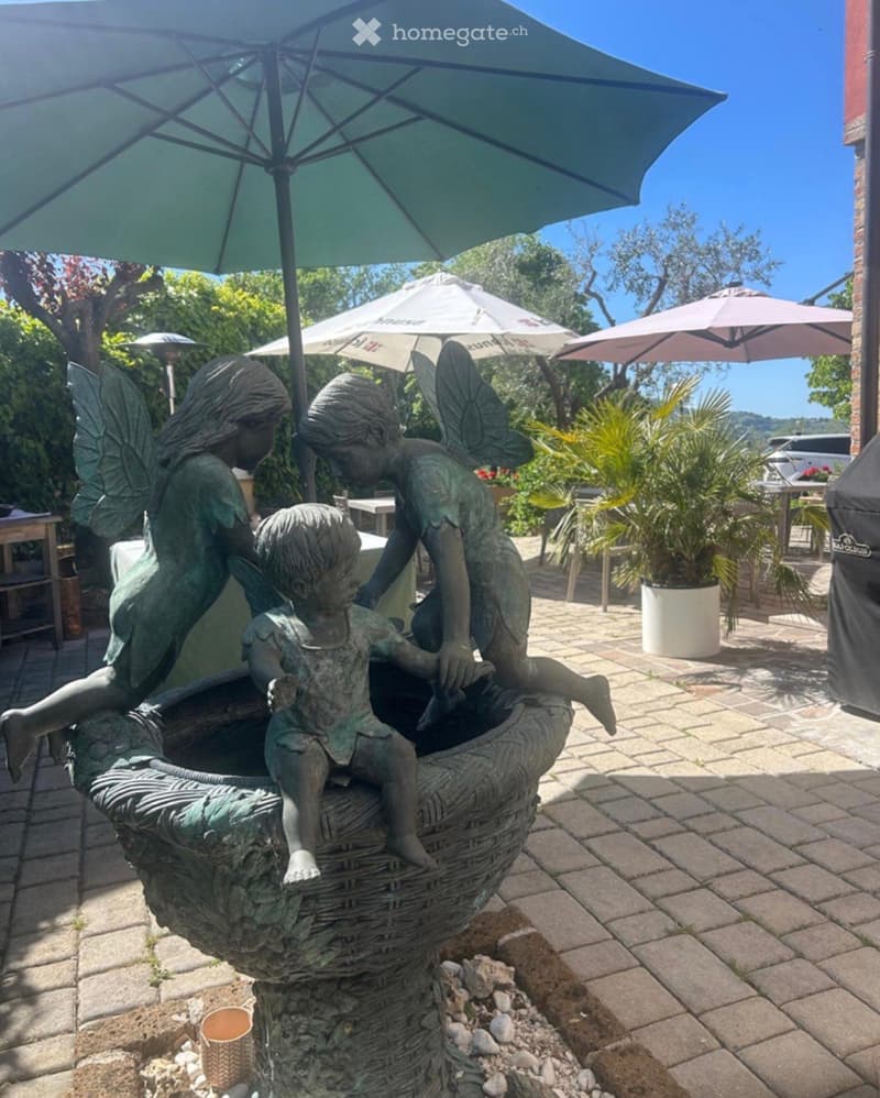 Garden area with a decorative fountain featuring three cherub statues, surrounded by potted plants, outdoor tables, and umbrellas.
