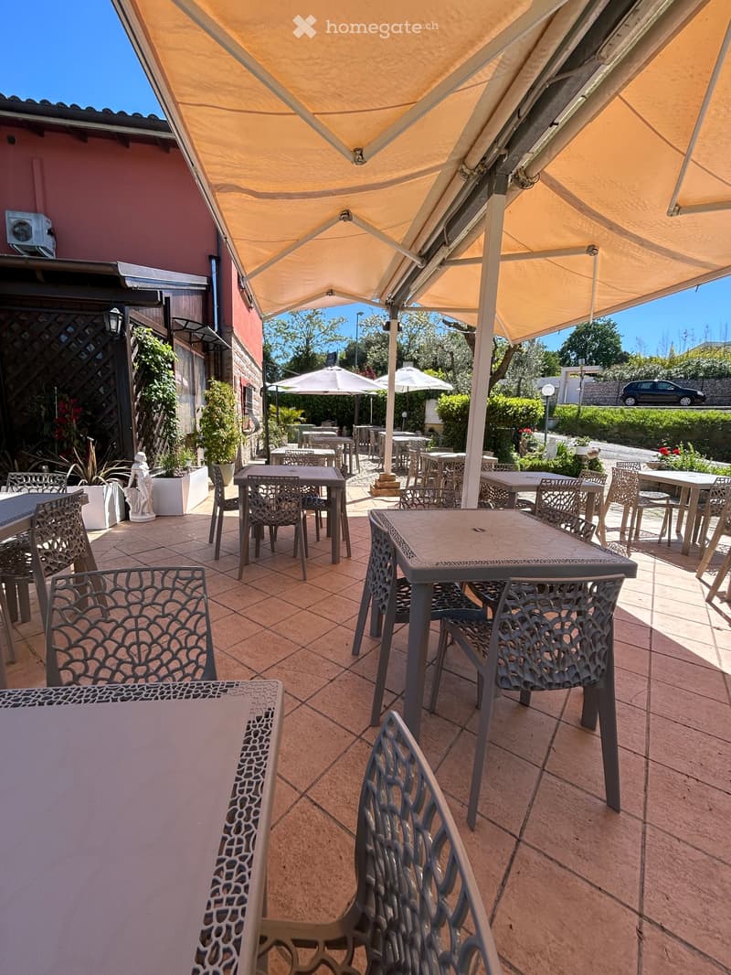 A covered outdoor seating area with tiled flooring, multiple tables and chairs arranged under a large canopy, with additional seating and umbrellas visible in the background.
