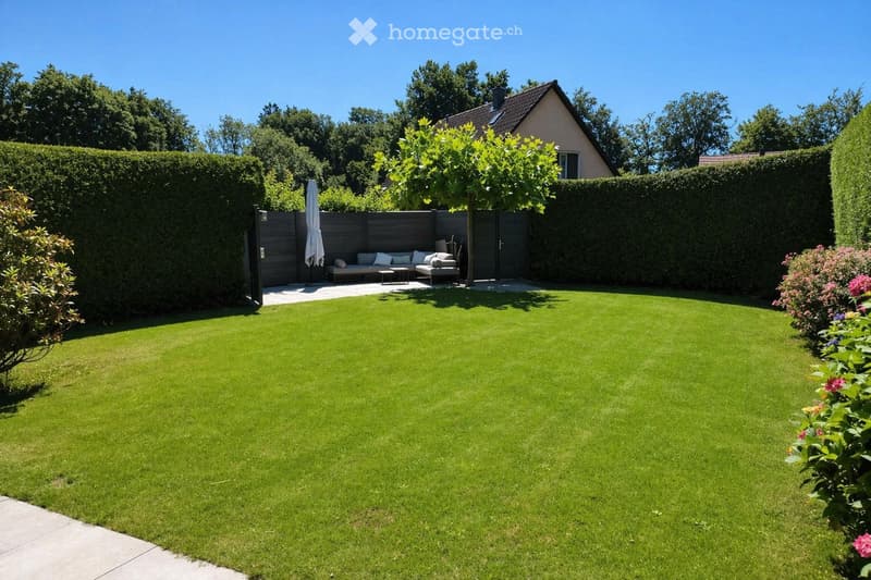 Well-maintained garden with a neatly trimmed lawn, a small seating area with a sofa and table, and a closed gate leading to the house.
