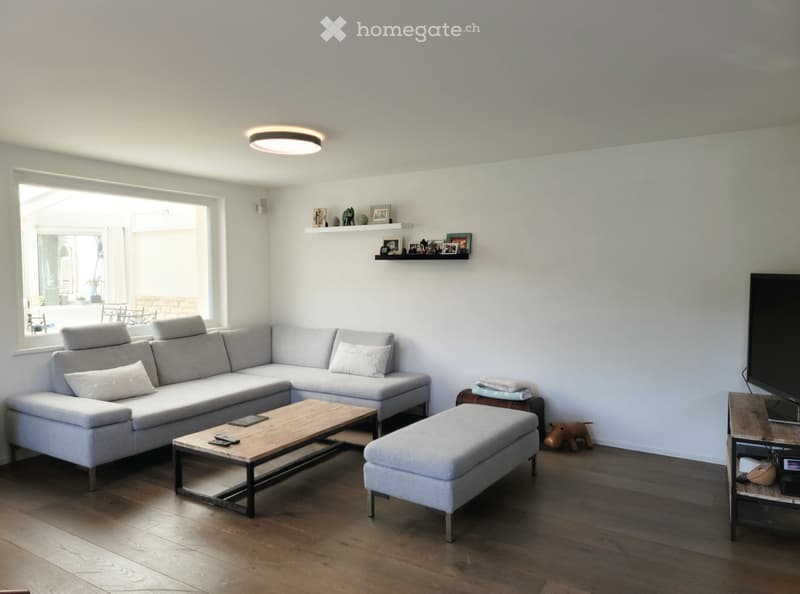 Modern living room with light grey sectional sofa, wooden coffee table, ottoman, TV stand, and decorative shelf with framed photos.
