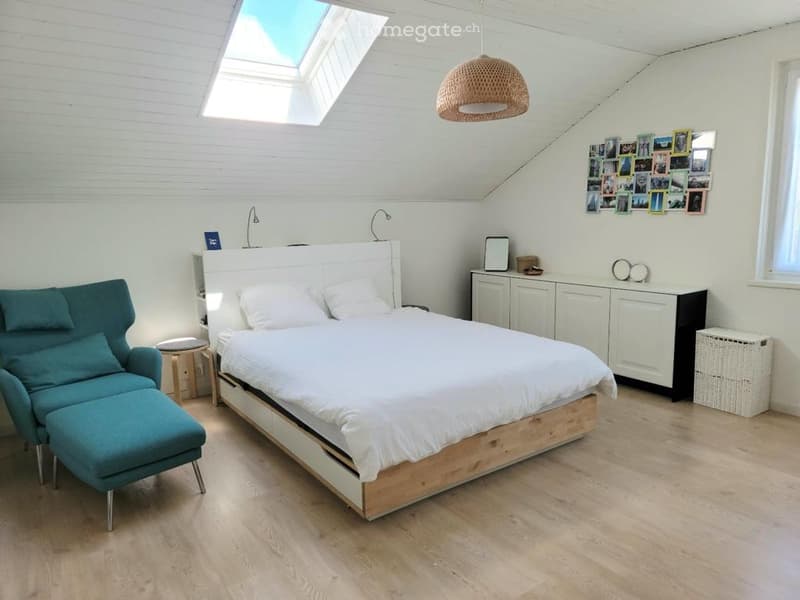 Bright, modern bedroom with a large bed, teal sofa, wooden stool, white cabinet, and a skylight window. The room features a slanted ceiling and a cozy, inviting atmosphere.