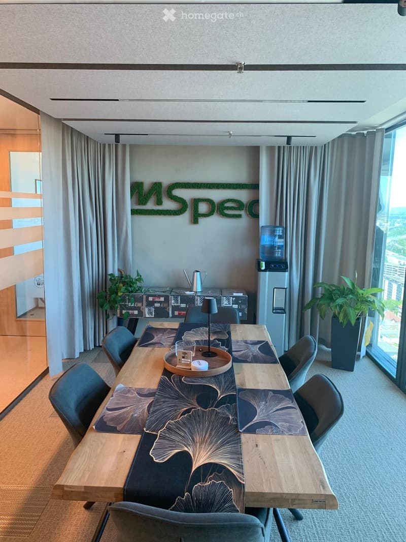 Modern conference room with a long wooden table, chairs, a lamp, a water cooler, and plants. The room features large windows with a city view and a decorative sign on the wall.