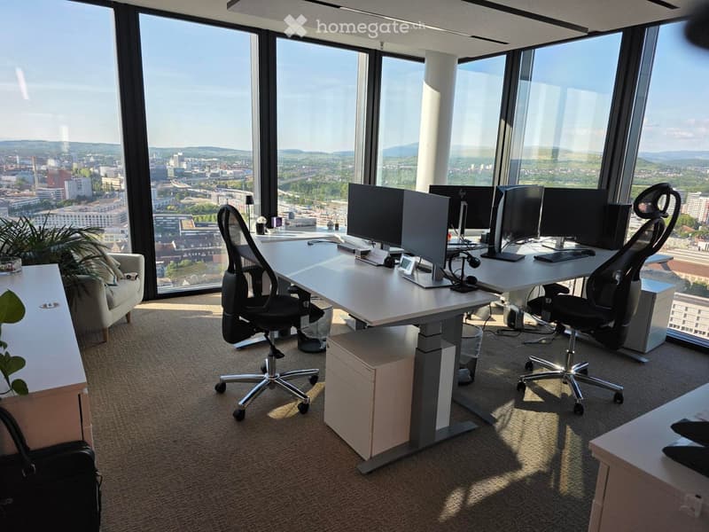 Modern office space with large windows offering a city view, equipped with desks, chairs, multiple screens, and a small sofa.
