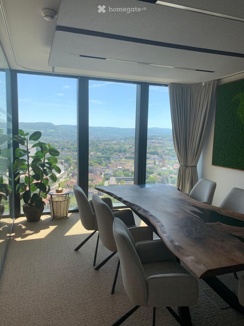 Bright room with large windows offering a scenic view, furnished with a long wooden table, several chairs, a potted plant, and curtains.