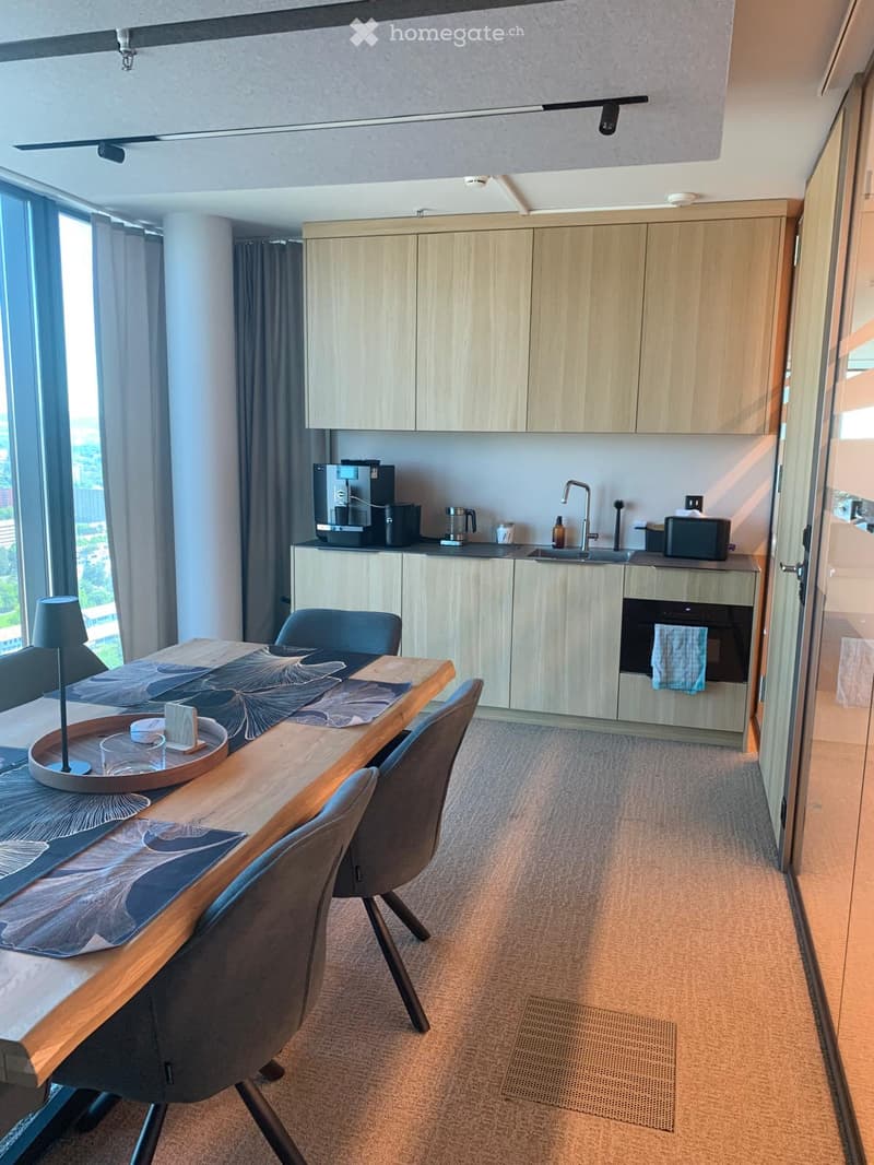 Modern kitchen and dining area with light wood cabinets, stainless steel appliances, a long wooden dining table with chairs, and a lamp on the table.
