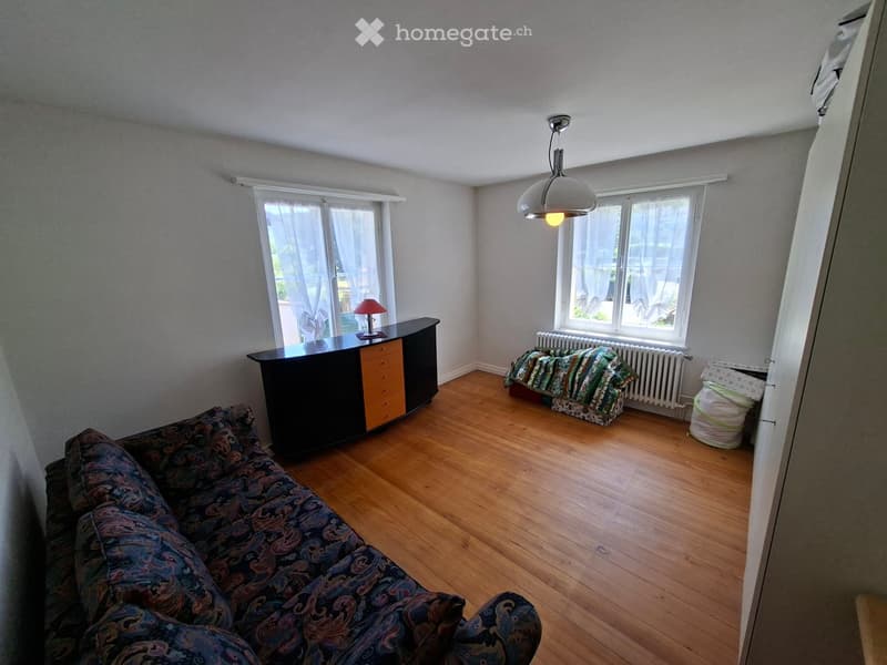 Living room with wooden flooring, large windows, sofa, table, lamp, and bed visible.