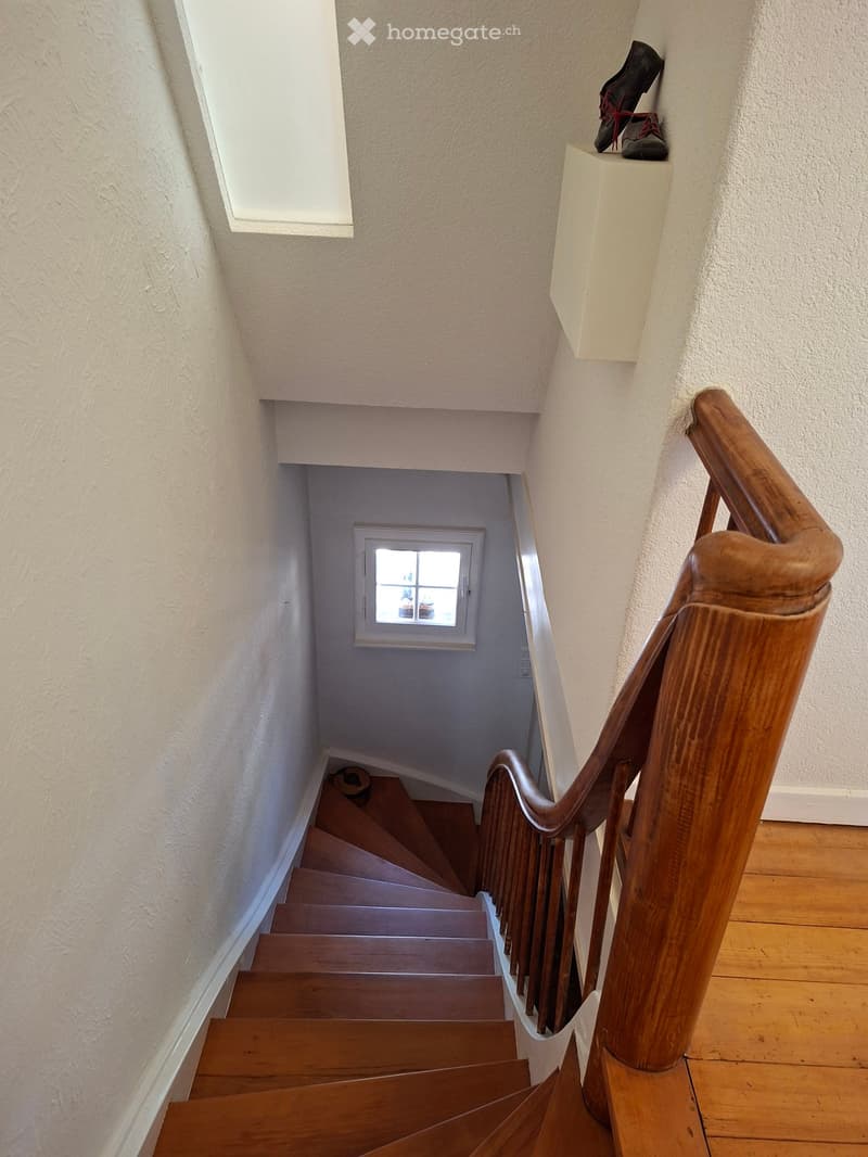 Wooden staircase with a wooden handrail, beige walls, and a small window at the top of the stairs.