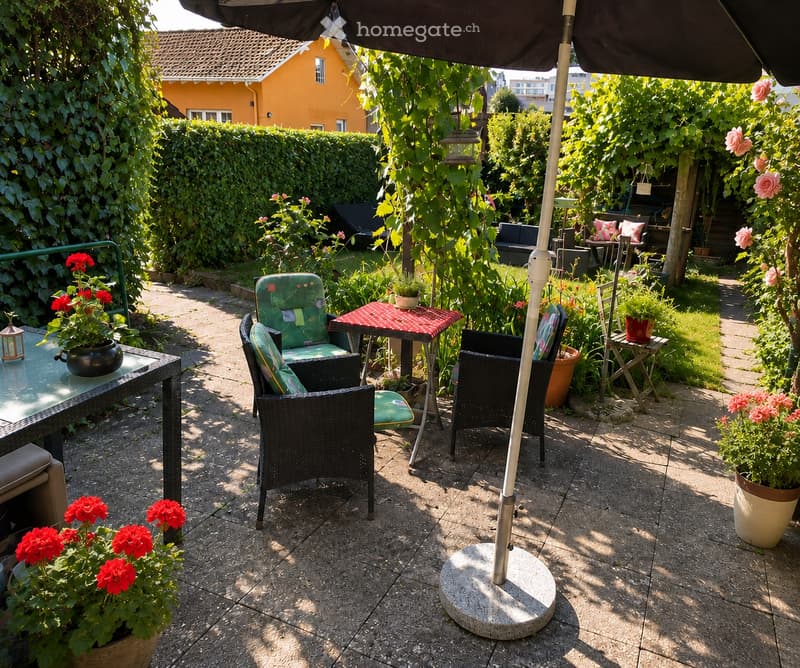 Garden patio with a glass table, wicker chairs, umbrella, and potted flowers. The area is surrounded by greenery and a hedge, with a yellow house visible in the background.