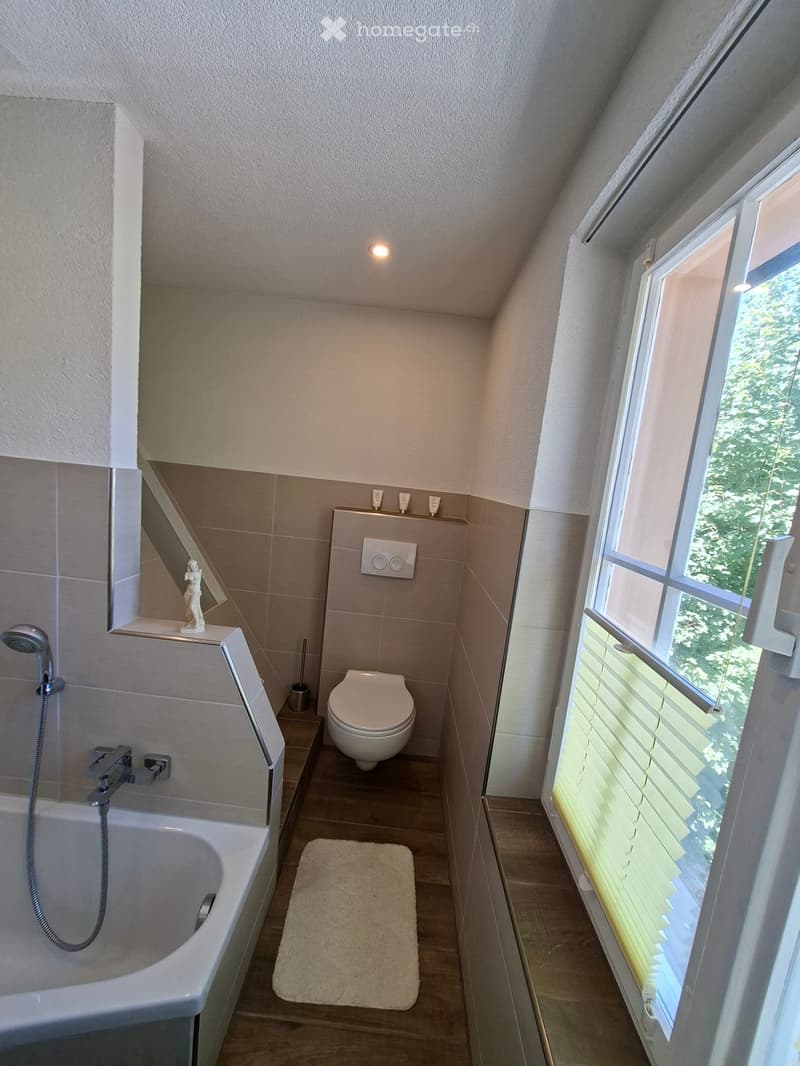 Modern bathroom with a bathtub, toilet, showerhead, and a small window with blinds. Includes a bathroom rug and a light switch on the wall.