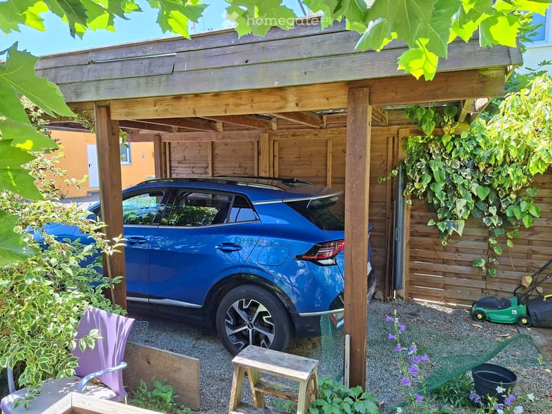 Wooden carport with a blue SUV parked underneath, surrounded by greenery, a small wooden stool, a purple folding chair, and a green lawnmower.