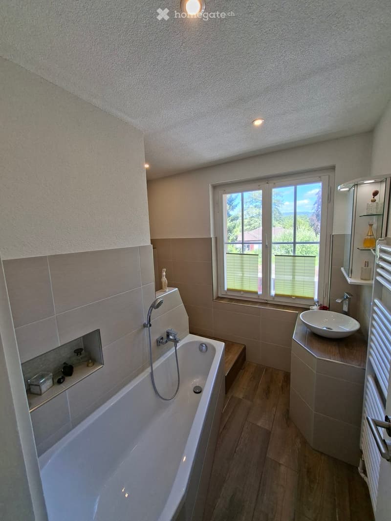 Modern bathroom with a bathtub, sink, mirror, and radiator. The room features large windows with blinds and a view of greenery outside.