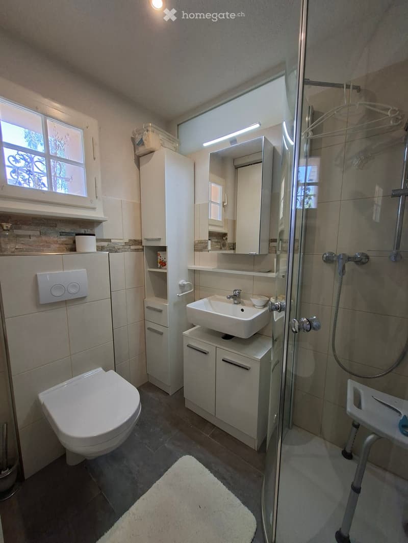 Modern bathroom with a toilet, sink, mirror, and glass-enclosed shower. Features white cabinets, a window, and a small white rug on the floor.