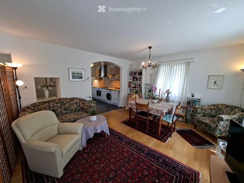 A spacious living room with a floral-patterned sofa, a cream-colored armchair, a small round table with a plant, a dining area with a table and chairs, a bookshelf, and a modern kitchen area with an oven and washing machine.