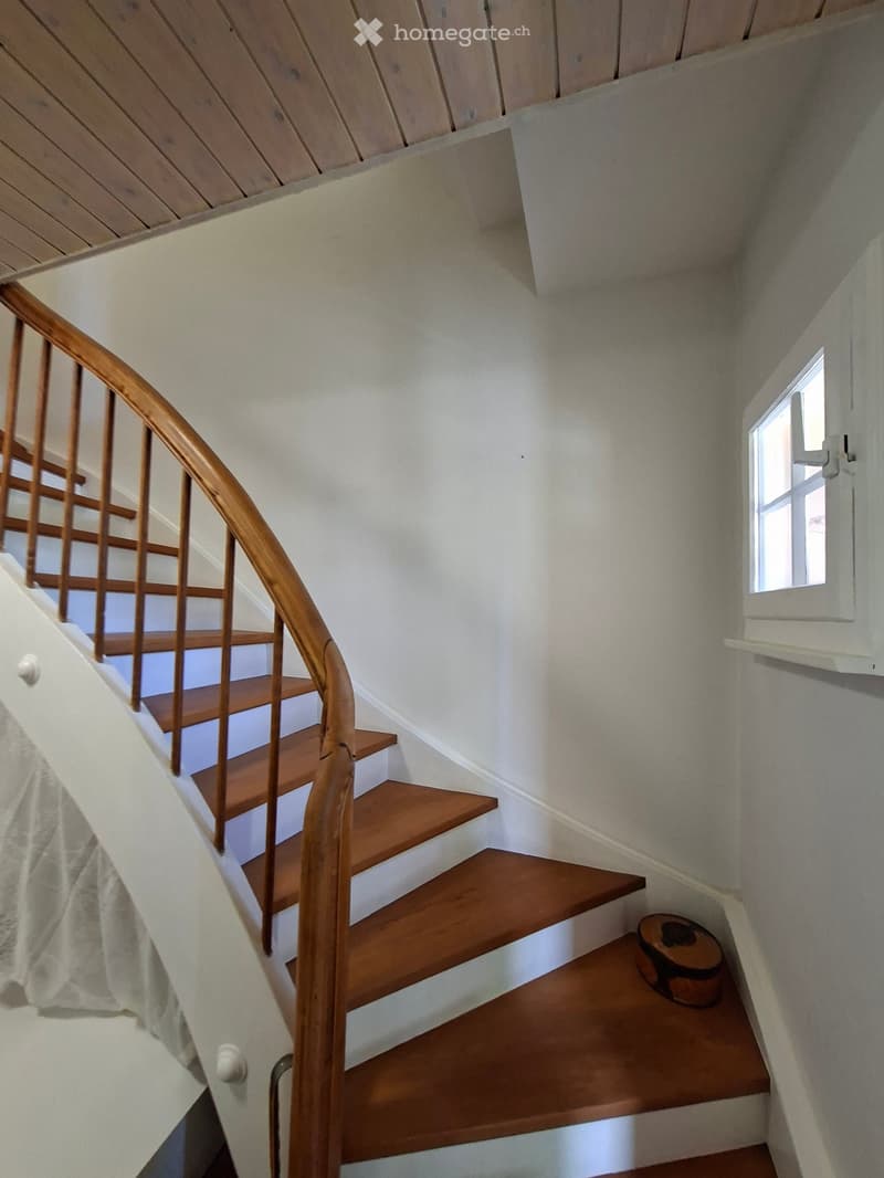 Wooden staircase with a curved wooden handrail, white walls, and a small window on the right side.
