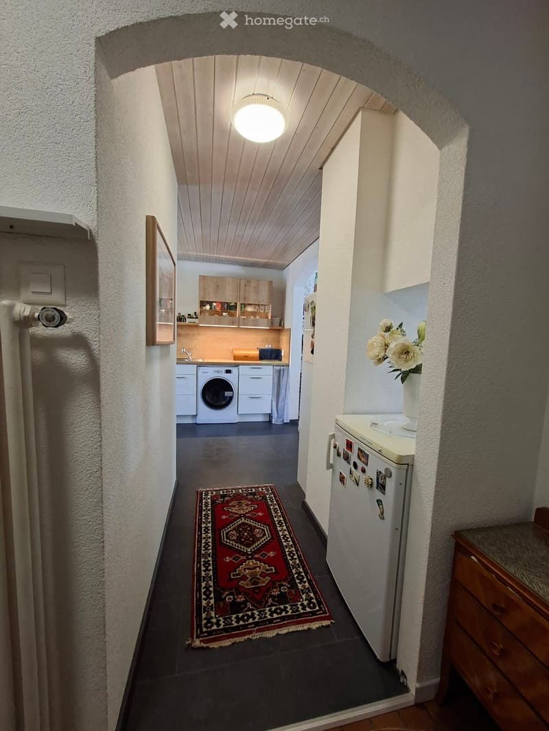A narrow hallway with a patterned red runner rug, leading to a kitchen area with white cabinets, a washing machine, and a refrigerator with magnets. The hallway has textured white walls and a wooden ceiling.
