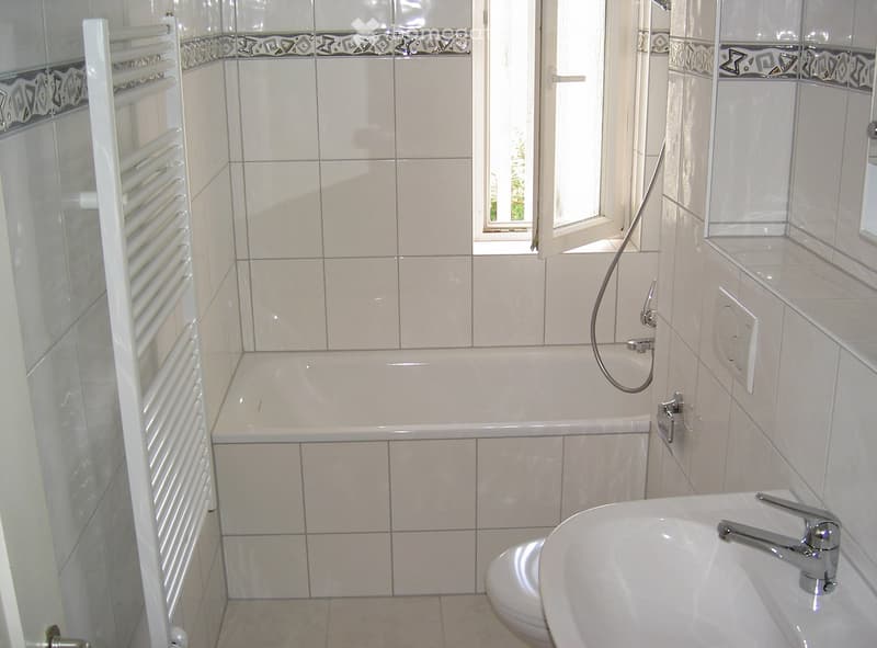 Bathroom with a bathtub, sink, toilet, and a radiator. The walls are tiled with a decorative border, and there is a window with a view of greenery outside.