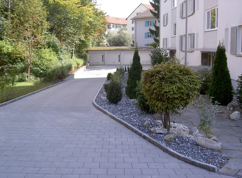 Brick driveway leading to a garage with surrounding greenery and decorative rock garden.