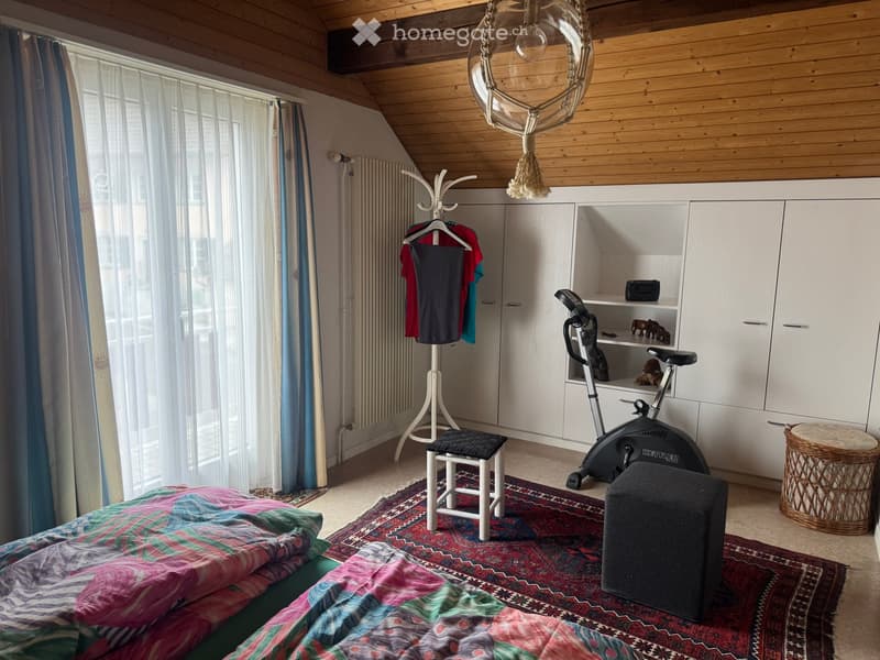 Cozy attic bedroom with wooden slanted ceilings, large window with curtains, colorful bedspread, exercise bike, white wardrobe, and decorative items.