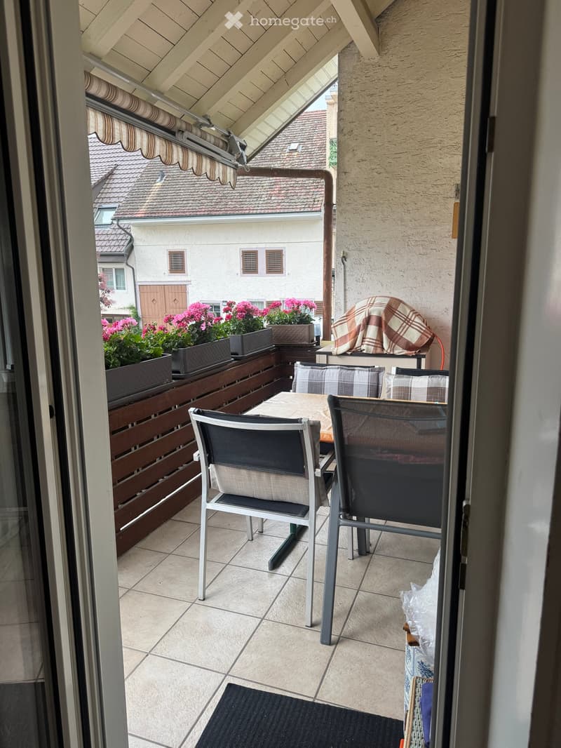 Balcony with tiled floor, wooden railing, and outdoor furniture including a table and two chairs, with several flower pots on the railing.
