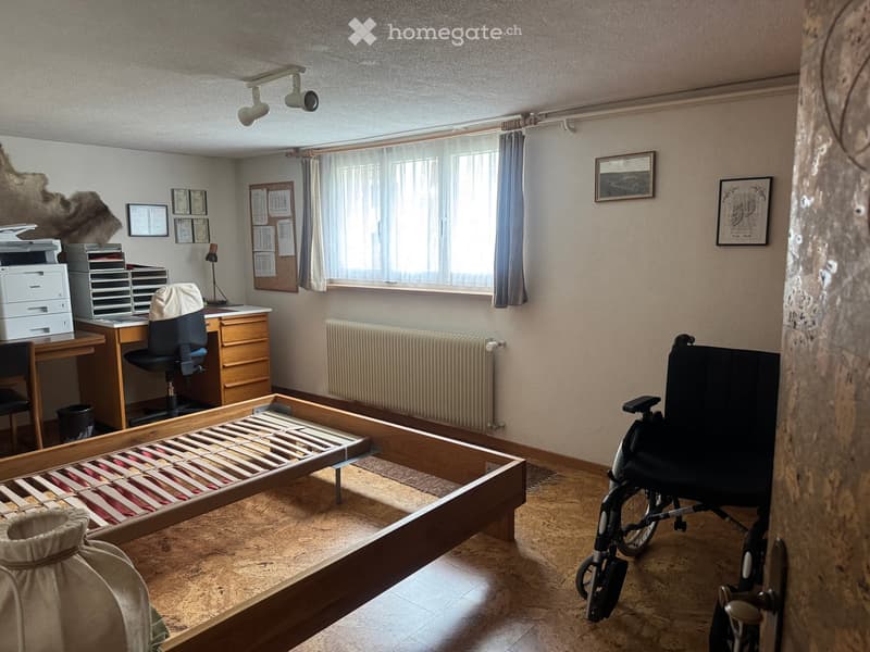 A furnished room with a wooden bed frame, a desk with a chair, a printer, a filing cabinet, a lamp, a radiator, a wheelchair, and a window with curtains.