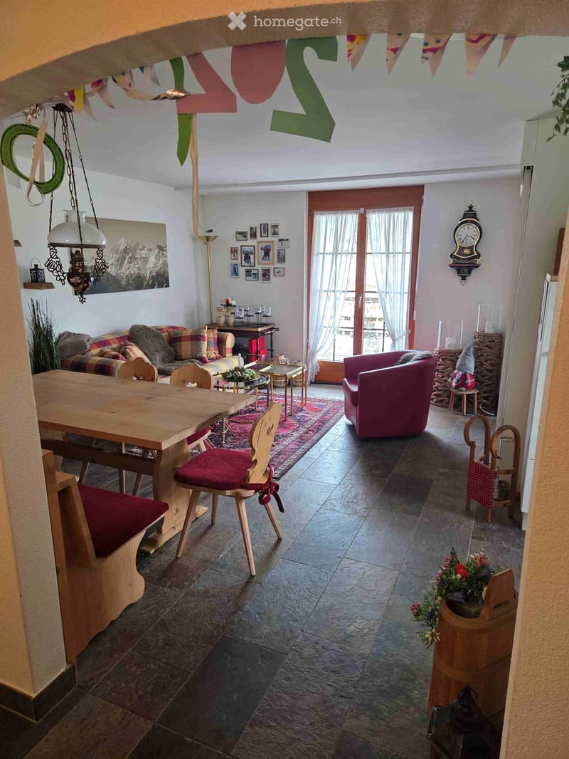 A cozy living and dining area with a wooden dining table, bench seating, a plaid-patterned sofa, a pink armchair, and a small coffee table. The space features a stone-tiled floor, a hanging chandelier, and a wall-mounted clock.