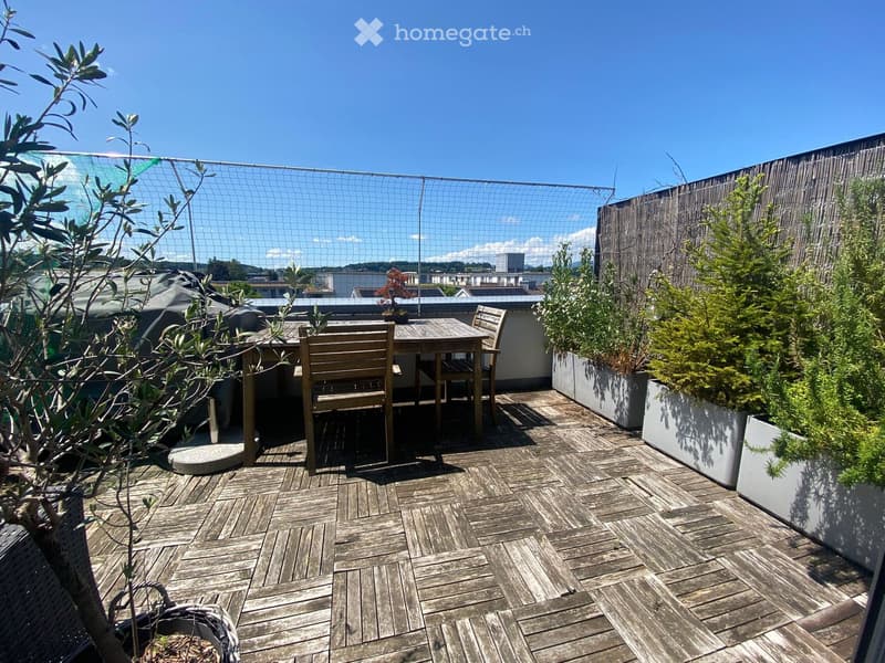 Wooden decked balcony with a small table and chairs, surrounded by potted plants and a view of the city.