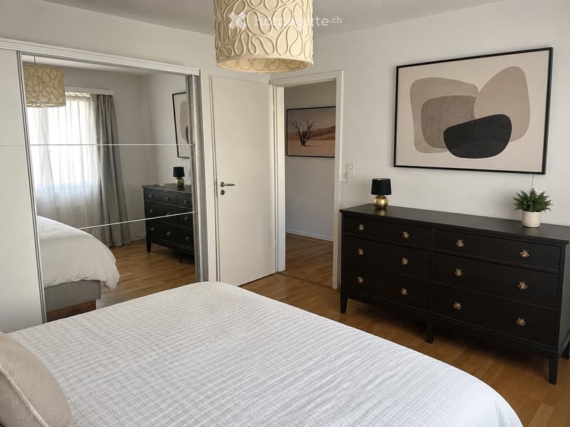 Modern bedroom with a large bed, nightstand, dresser, mirror, and artwork on the wall. The room features wooden flooring and a hanging light fixture.