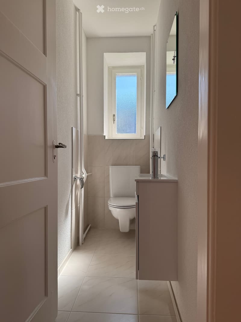 Small bathroom with white tiles, white toilet, white sink, mirror, window